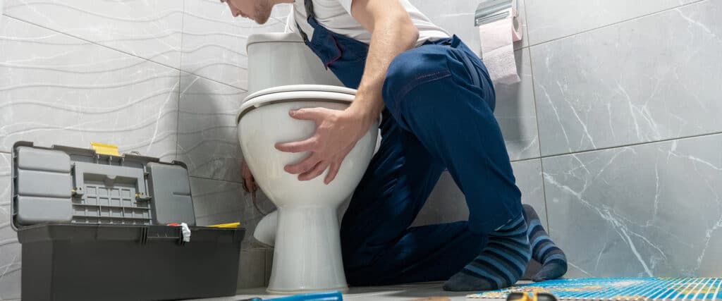 Plombier en salopette bleue réparant ou installant une toilette blanche dans une salle de bain carrelée, boîte à outils ouverte.