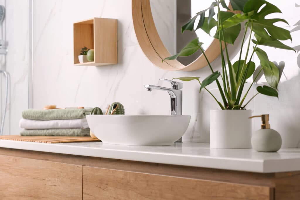 Salle de bain moderne et lumineuse avec meuble vasque en bois clair, évier blanc, miroir rond et plante Monstera. Serviettes vertes empilées.